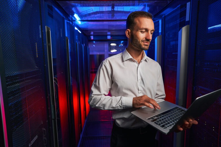 Caucasian programmer is looking to the screen of laptop while standing near the server rack